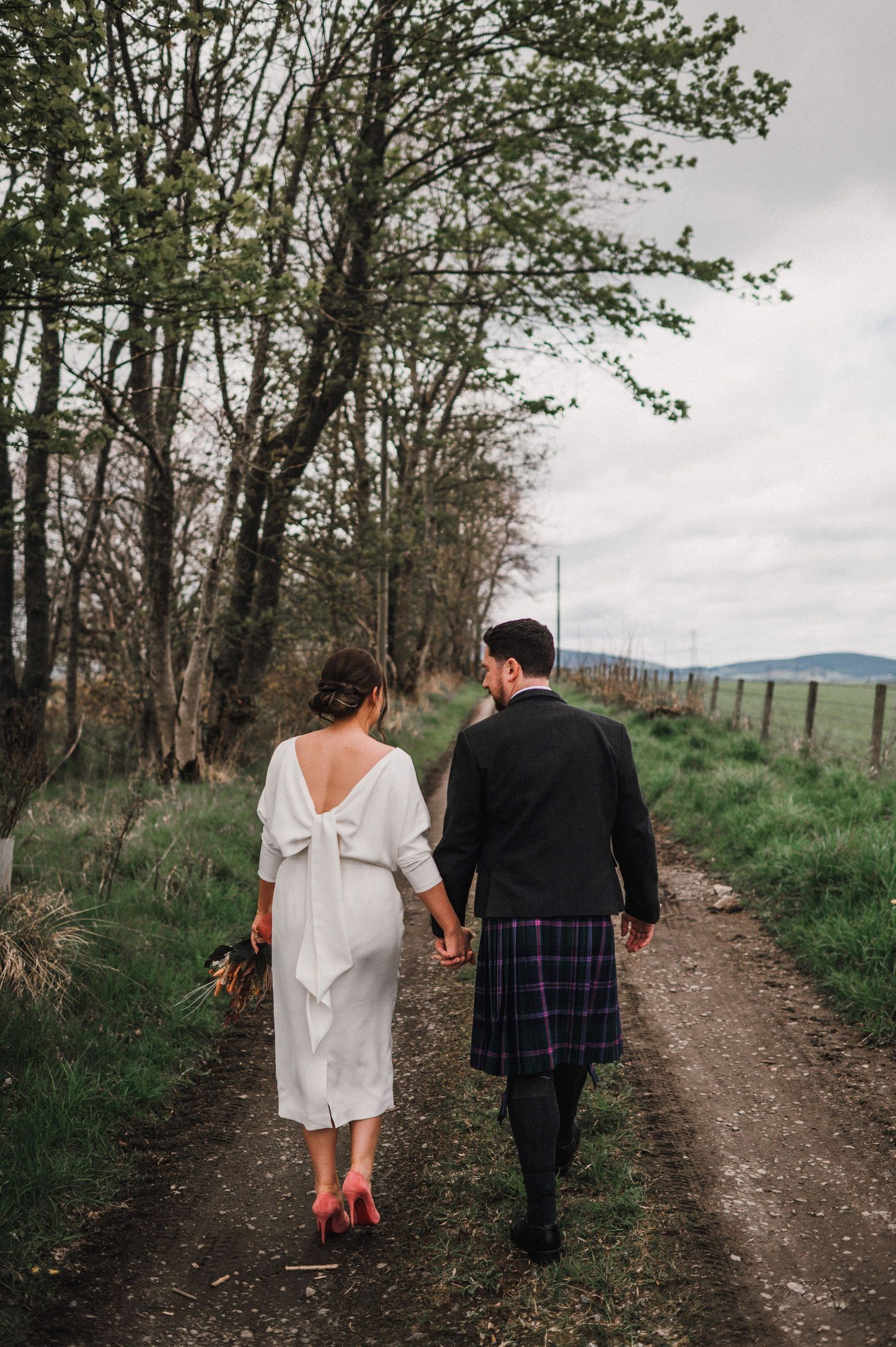 Couple walking hand in hand down a country path, wearing wedding attire with traditional Scottish kilt.