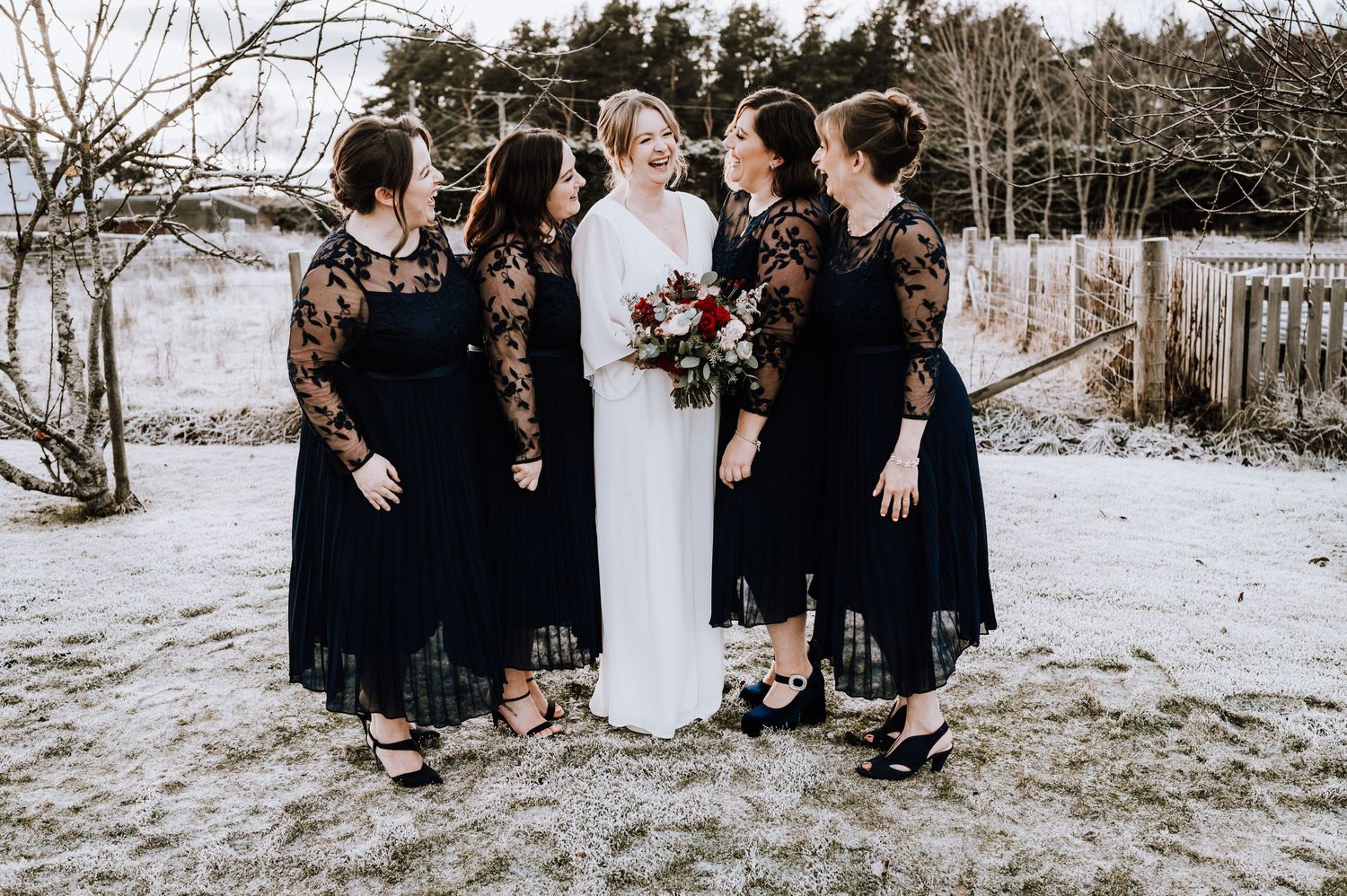 Same Sex Wedding Photography. Wedding party in black lace dresses poses with person in white gown against snowy backdrop.
