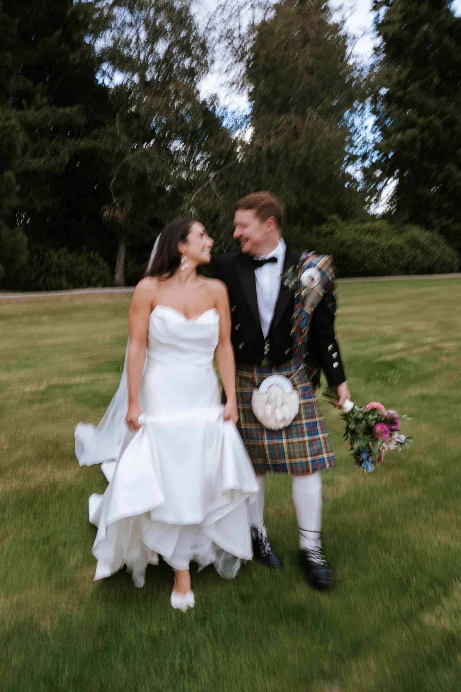 Romantic garden wedding photo with bride in flowing gown and groom in traditional Scottish kilt.