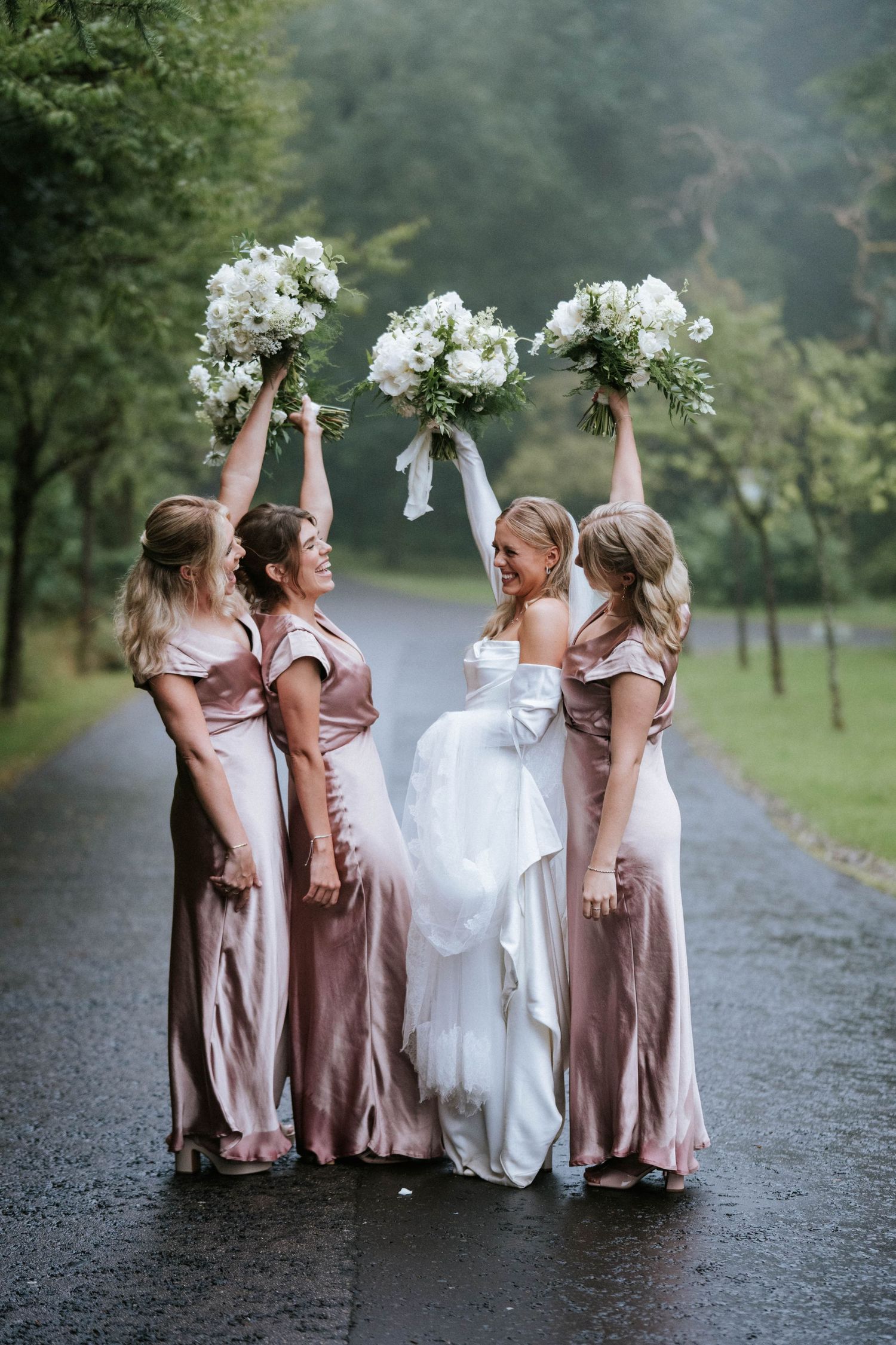 Bridesmaids in dusty rose dresses pose playfully on a rainy path.