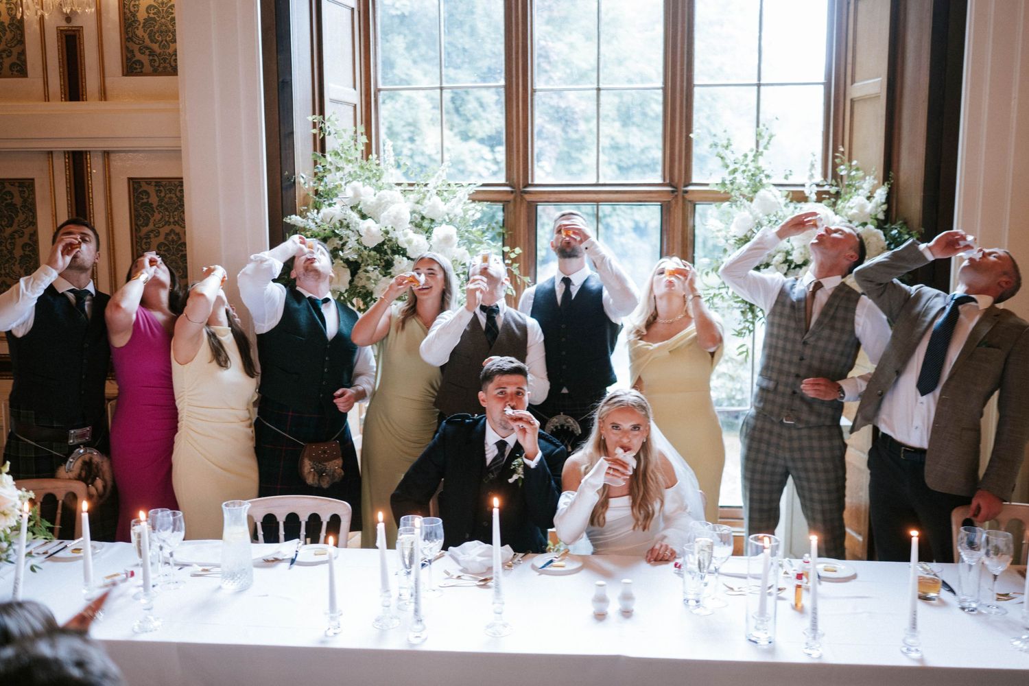 Wedding party guests pose together at a reception table with raised glasses in celebration.