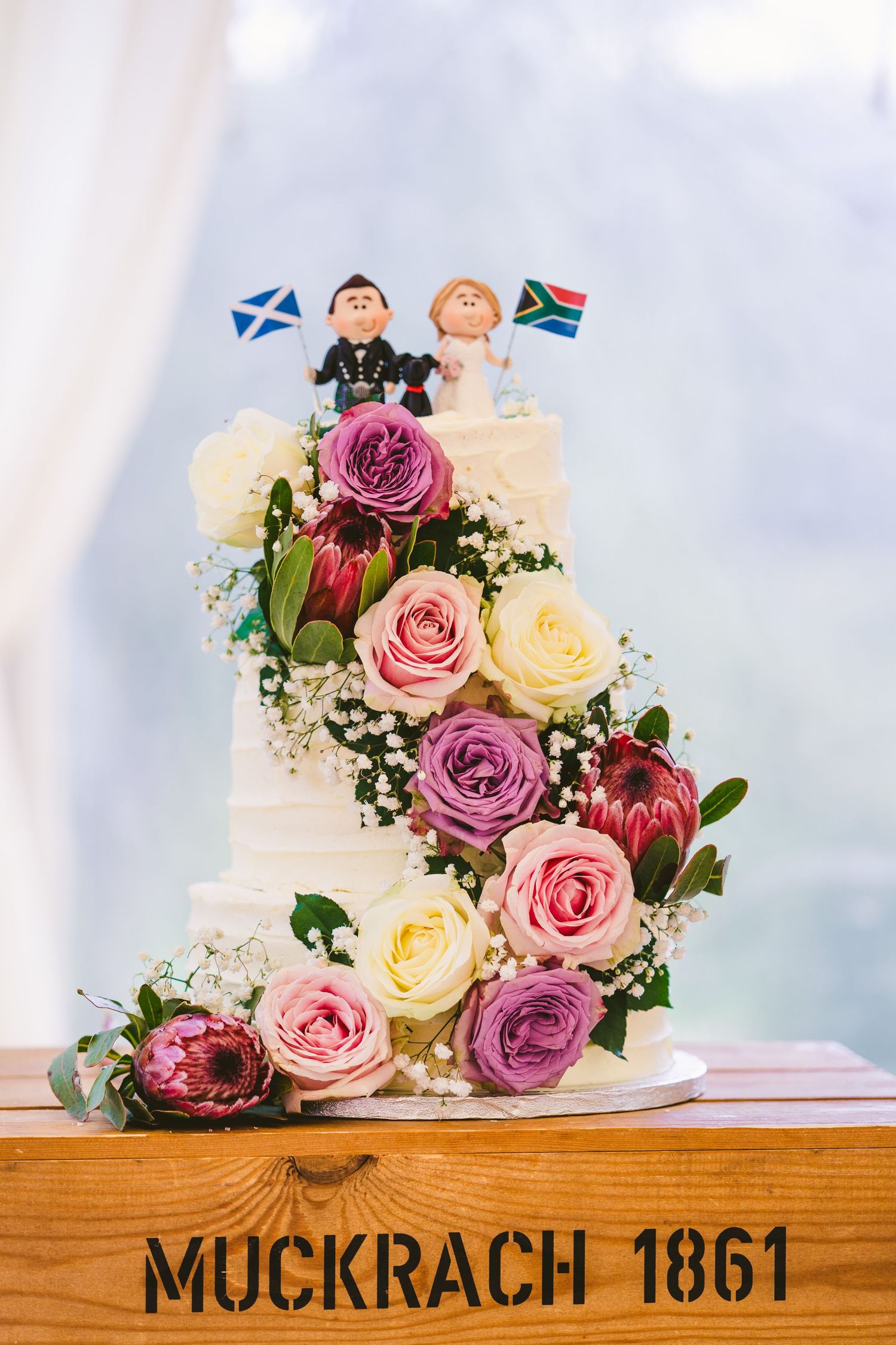 Wedding cake decorated with pink and purple roses sits on wooden surface marked Mockrach 1861.