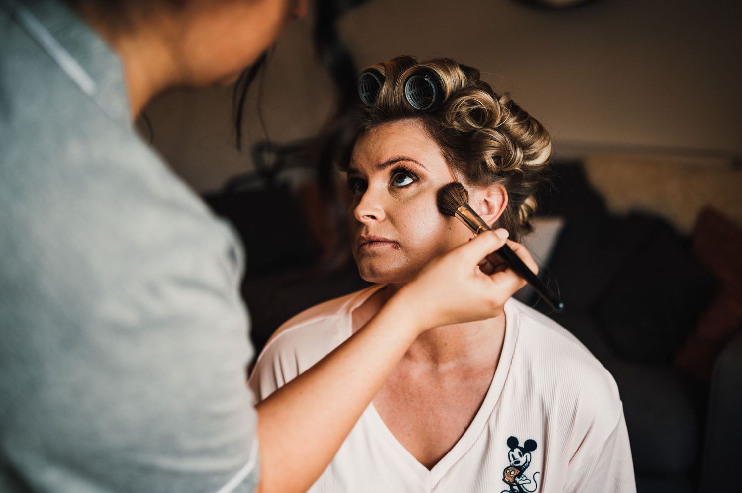 Professional makeup artist applies eyeshadow during bridal preparation in a dimly lit room.