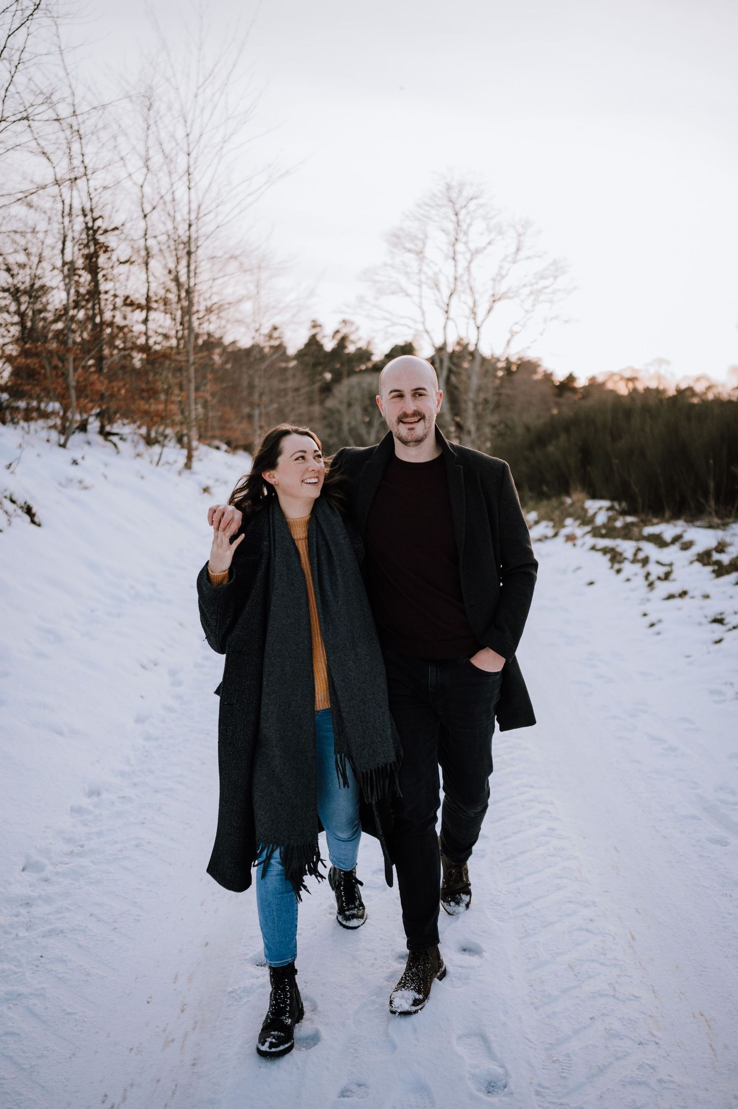 Two people walking together and laughing on a snowy winter path surrounded by bare trees at dusk.
