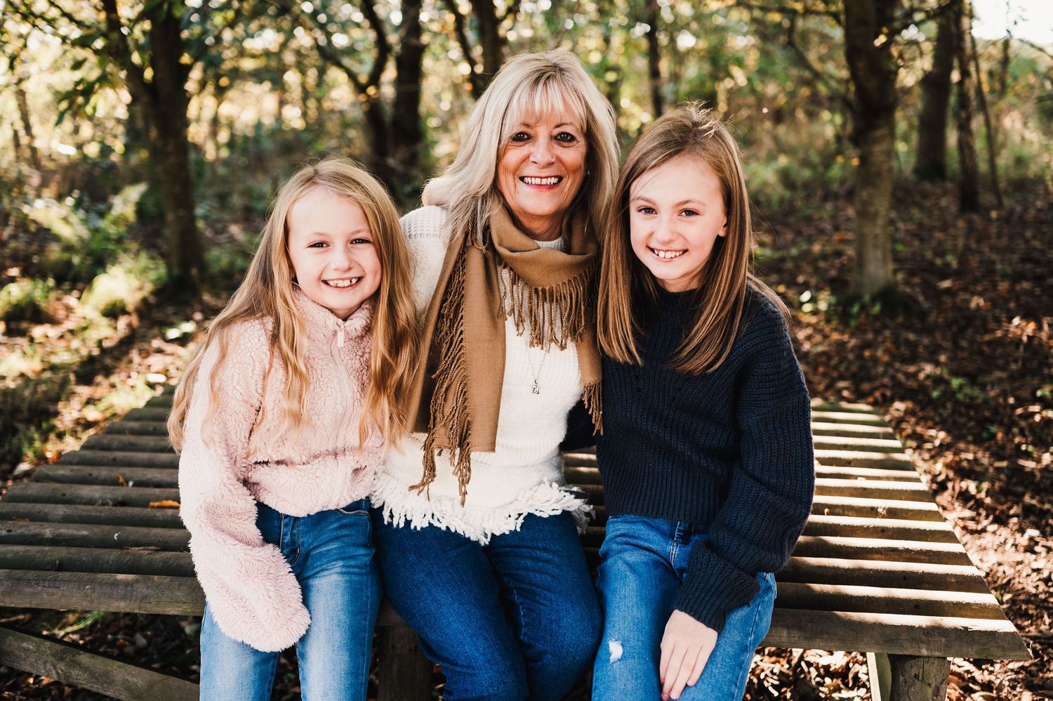 Three people sit together on wooden steps surrounded by autumn foliage in a natural outdoor setting.