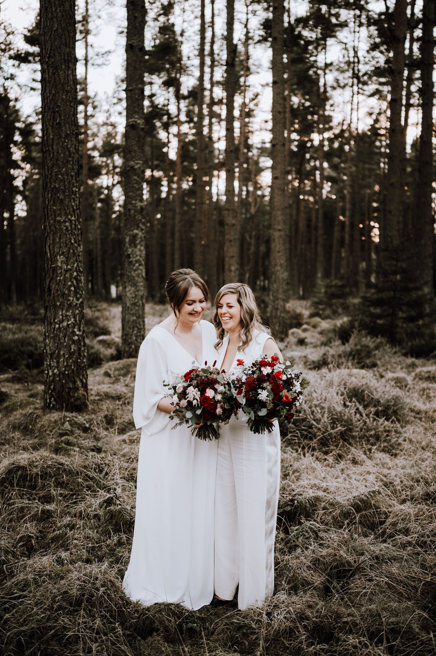 Same Sex Wedding Photography. Two people in white dresses hold deep red and burgundy bouquets while standing in a moody forest setting.