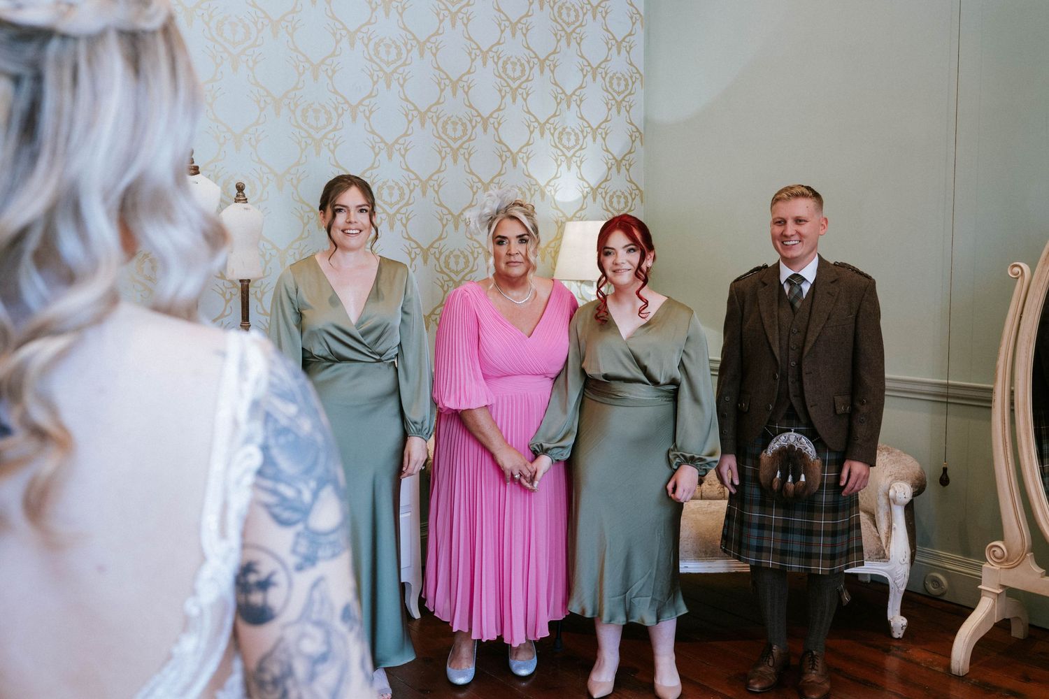 Wedding party members in pastel dresses pose with a person in traditional Scottish kilt.