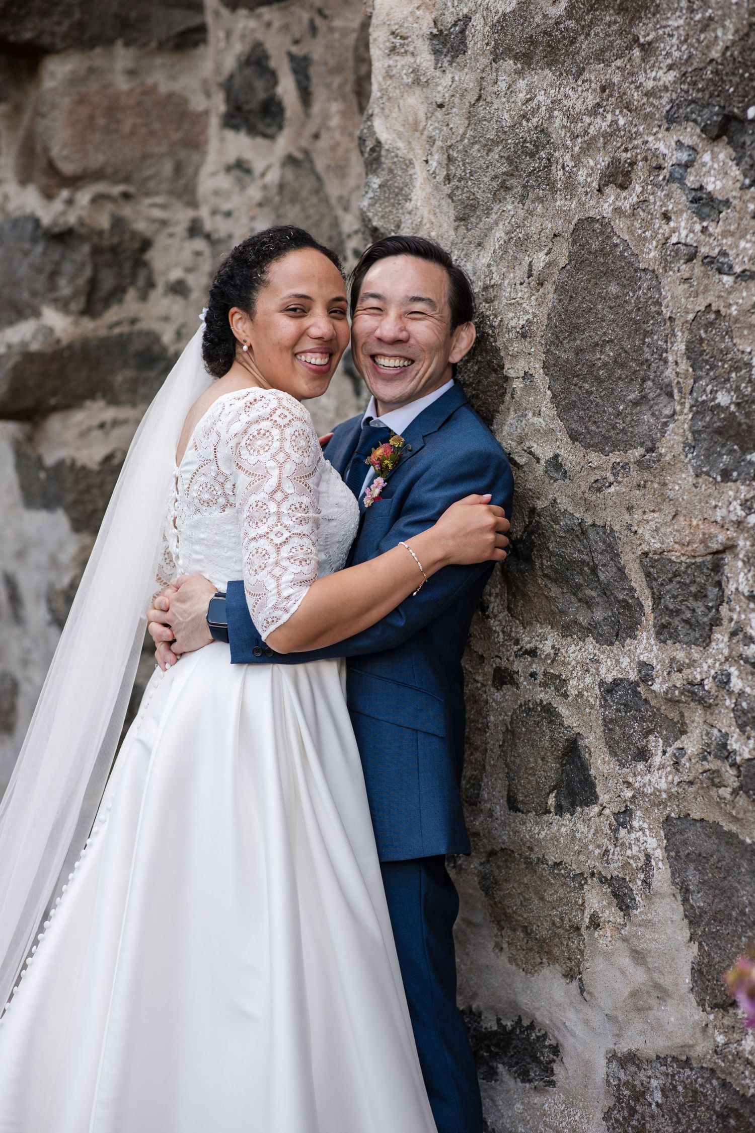 A joyful wedding couple embraces against a rustic stone wall, with the bride wearing a lace and white gown.