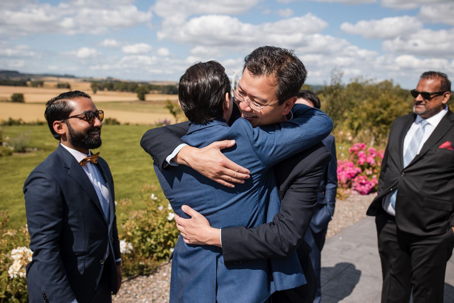 Wedding party members share emotional embraces outdoors against a scenic countryside backdrop.