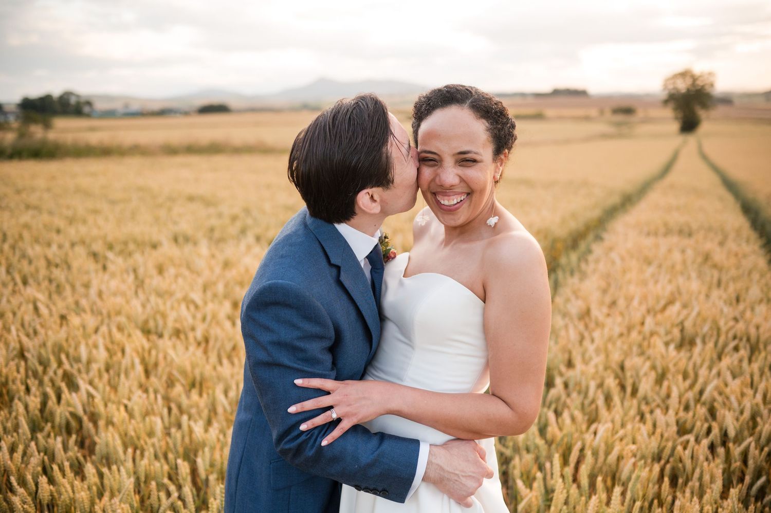Couple embracing in golden wheat field during sunset with countryside views.