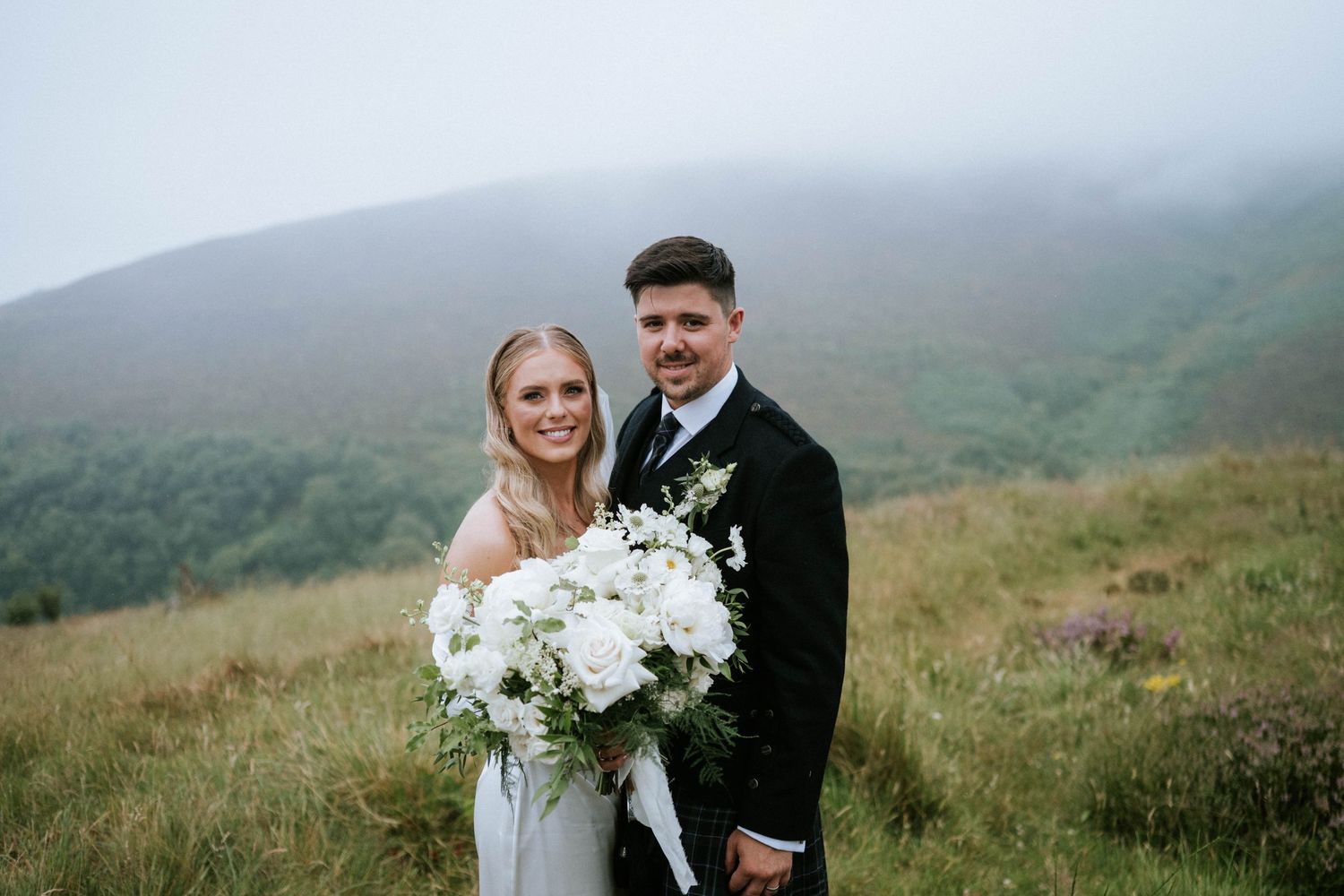Newlyweds pose together on misty green hillside holding white floral bouquet.