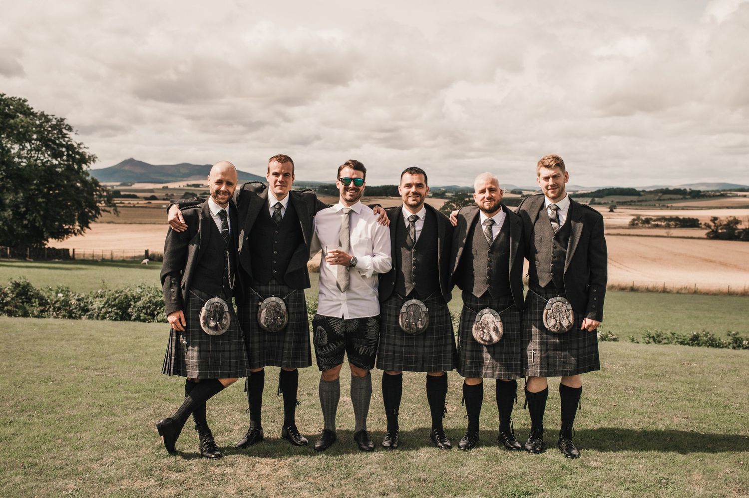 Groomsmen in traditional Scottish kilts stand together on a hilltop overlooking scenic countryside.
