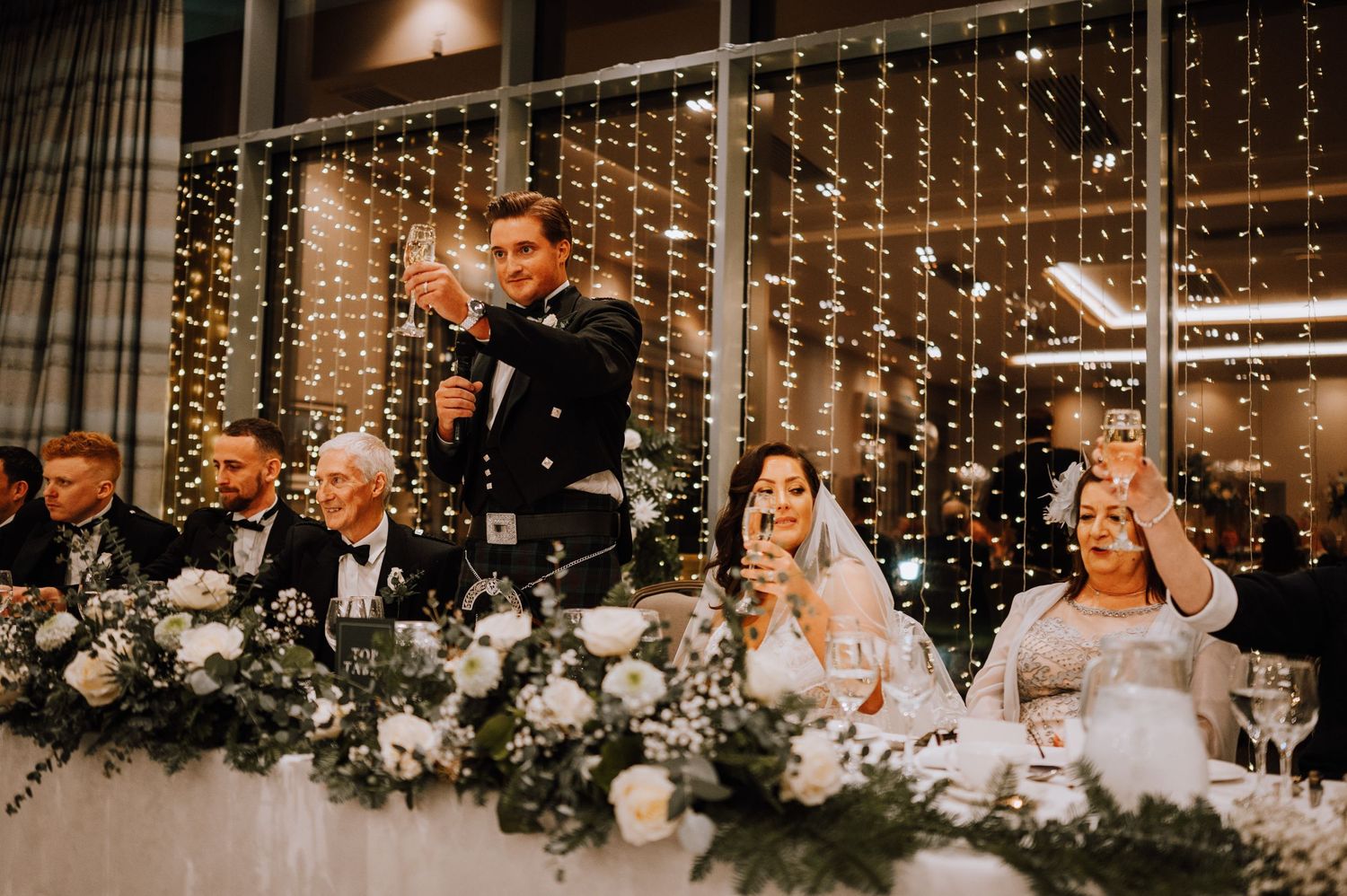 Wedding party seated at head table with elegant floral arrangements and twinkling light backdrop.