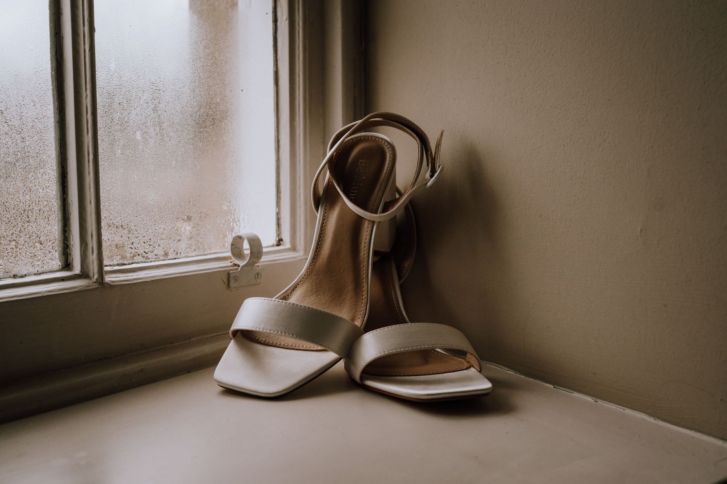 Elegant white wedding shoes sit on a windowsill in soft natural light.