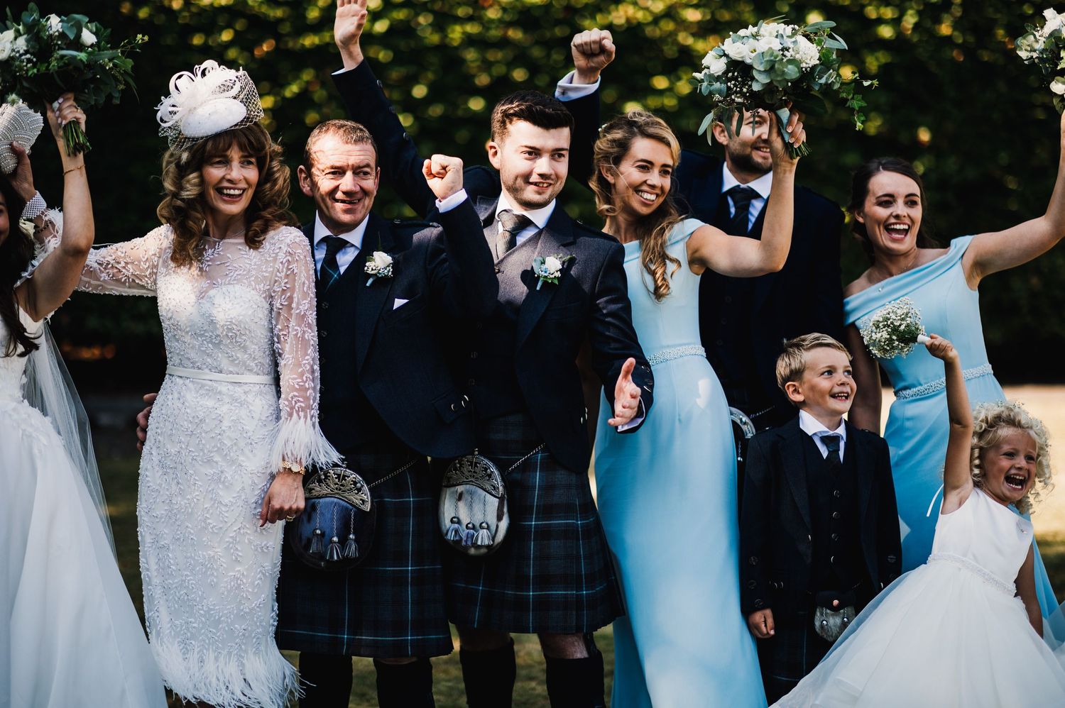 Joyful wedding group in Scottish formal wear celebrating with raised arms against garden backdrop.