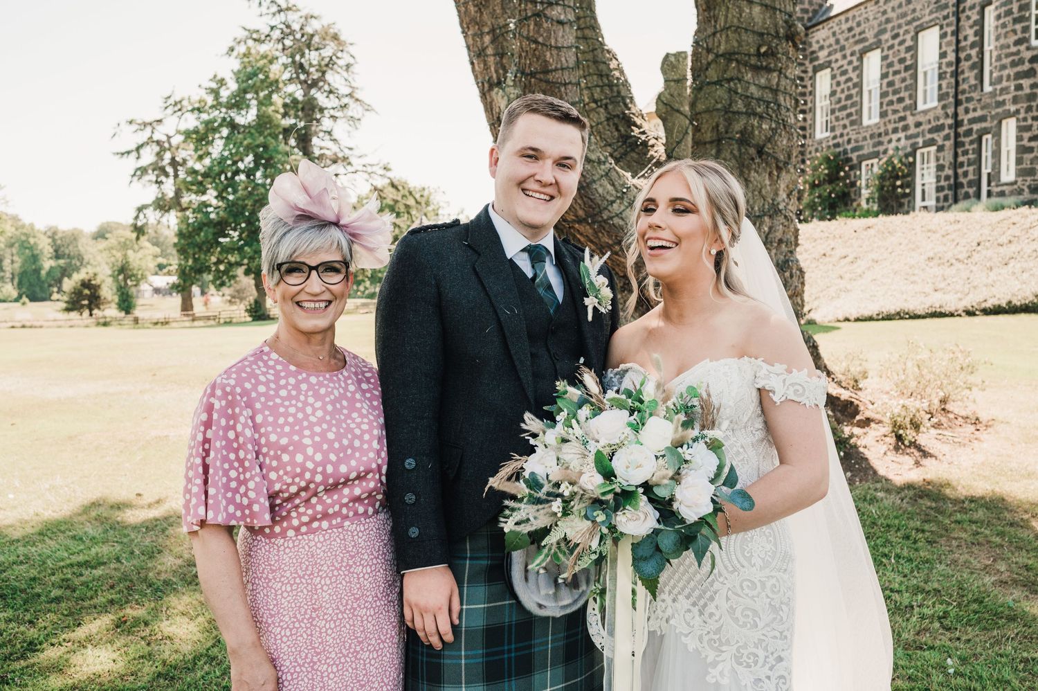 A wedding photo of a couple with a guest in pink outside a stone building.