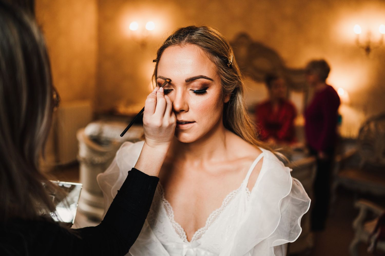Makeup application in progress during wedding preparations in a warmly lit room.