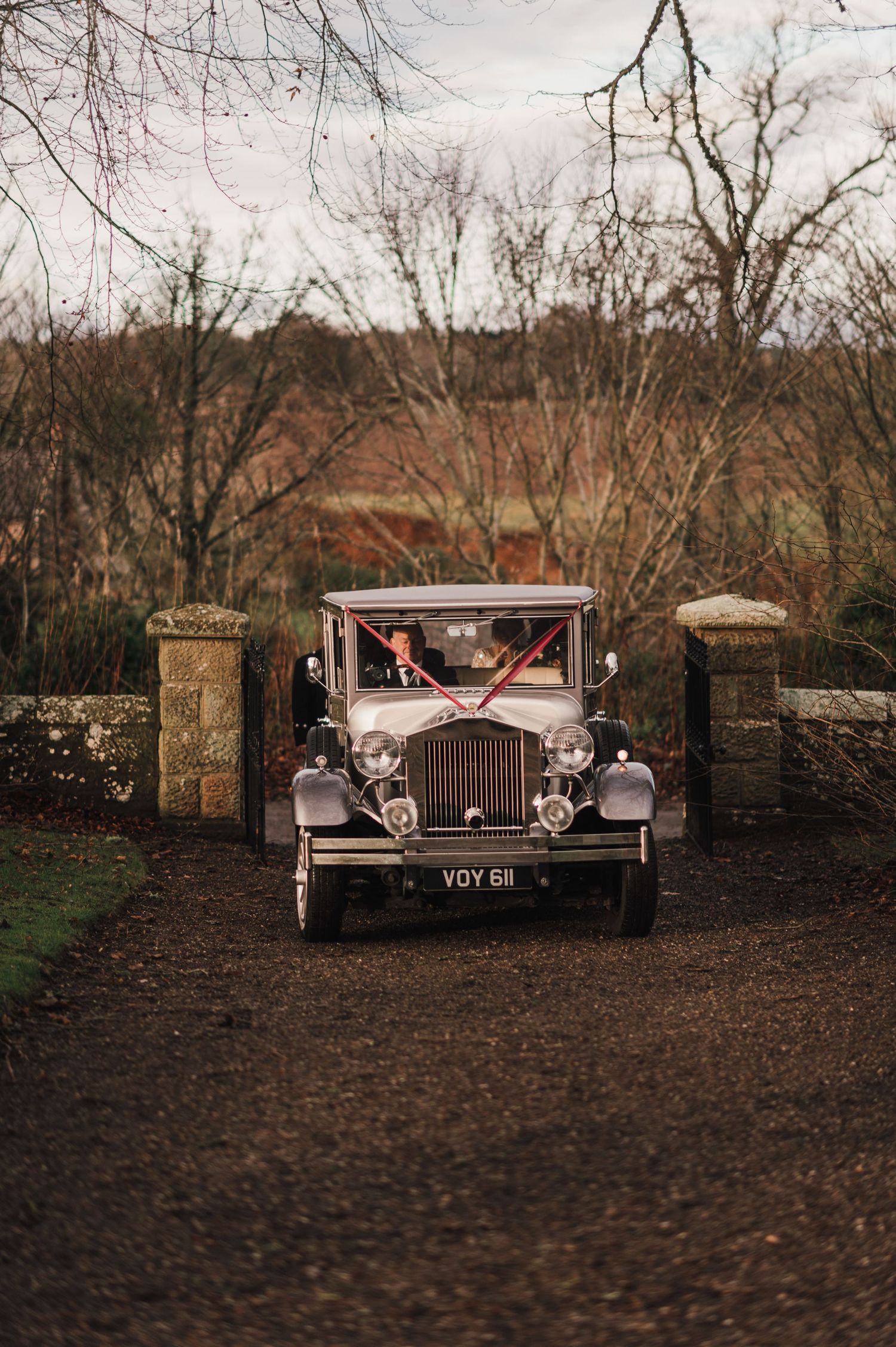 Vintage cream-colored wedding car parked between stone pillars on dirt path.