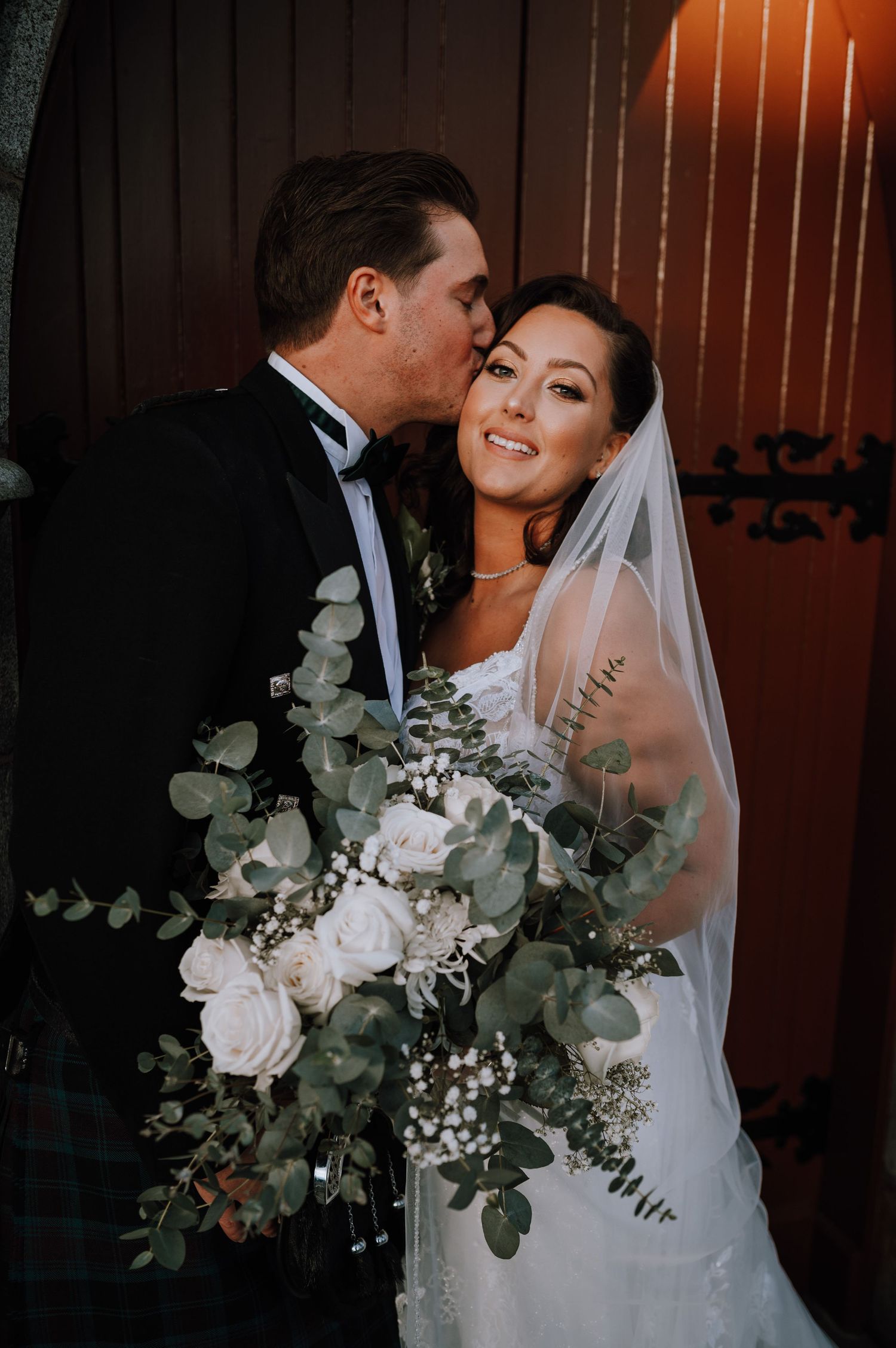 A romantic wedding portrait of a couple with a stunning bouquet of white roses and eucalyptus in dramatic lighting.