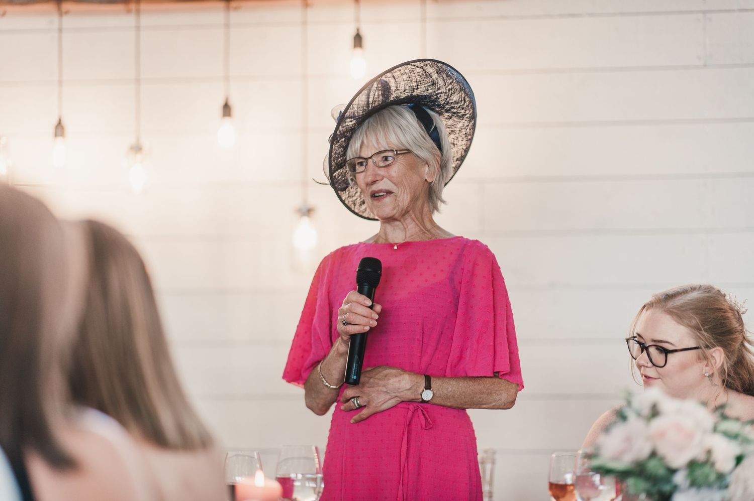 Wedding guest gives speech wearing pink dress and decorative hat at reception.
