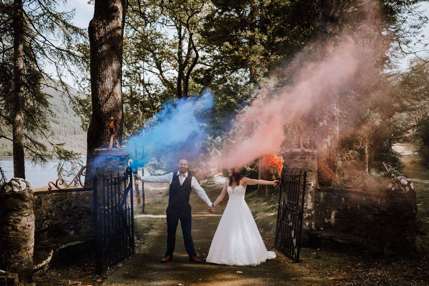 A couple celebrates with colorful smoke bombs in pink and blue at a garden gate entrance.