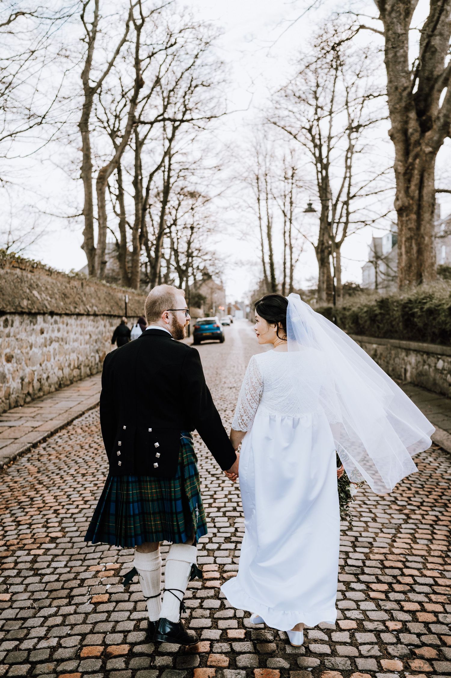 Newlyweds stroll down a cobblestone path, with the groom in a kilt and bride in a flowing white dress.