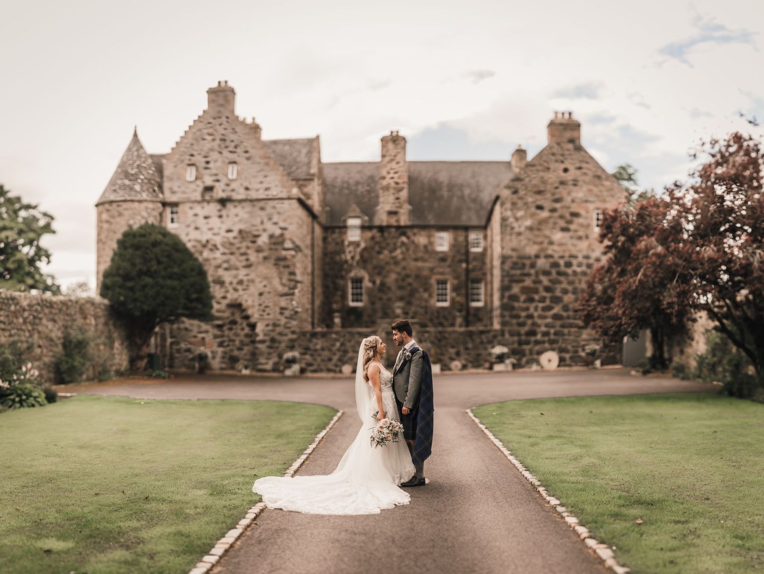 Newlyweds pose on pathway leading to stunning Scottish stone castle on their wedding day.