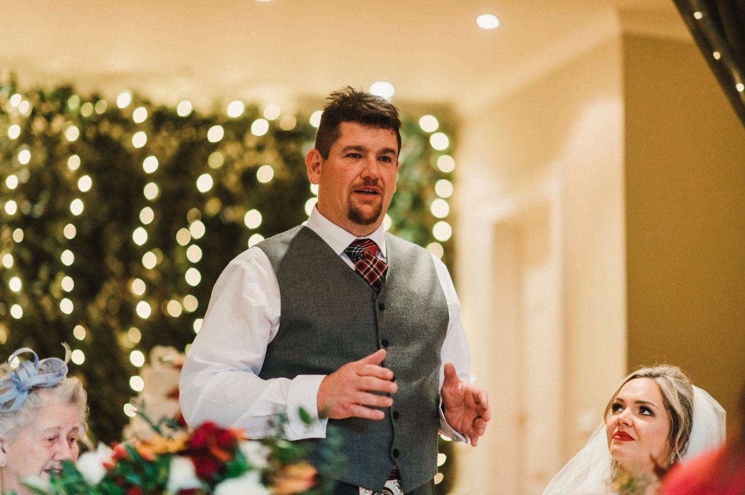 A wedding reception speech moment with festive string lights and floral centerpieces decorating the venue.