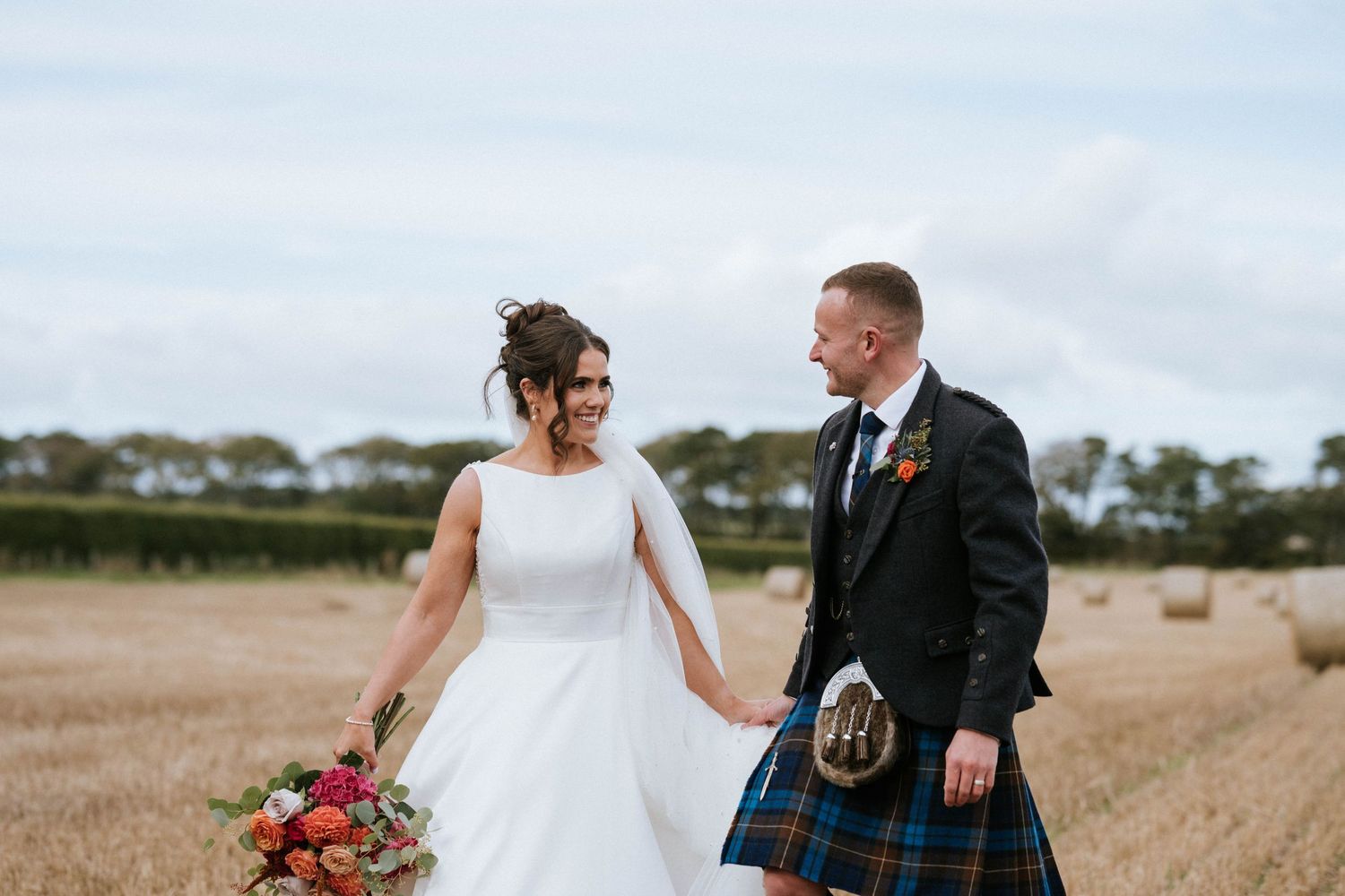 A couple shares a romantic moment in a harvest field, with the groom wearing traditional Scottish attire.
