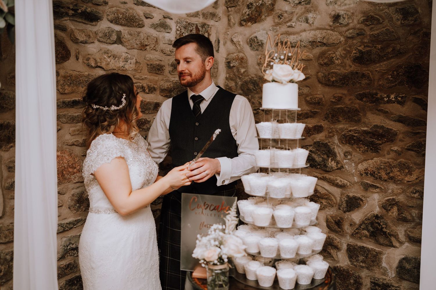 A romantic wedding scene with a tiered cupcake display against a stone wall backdrop.