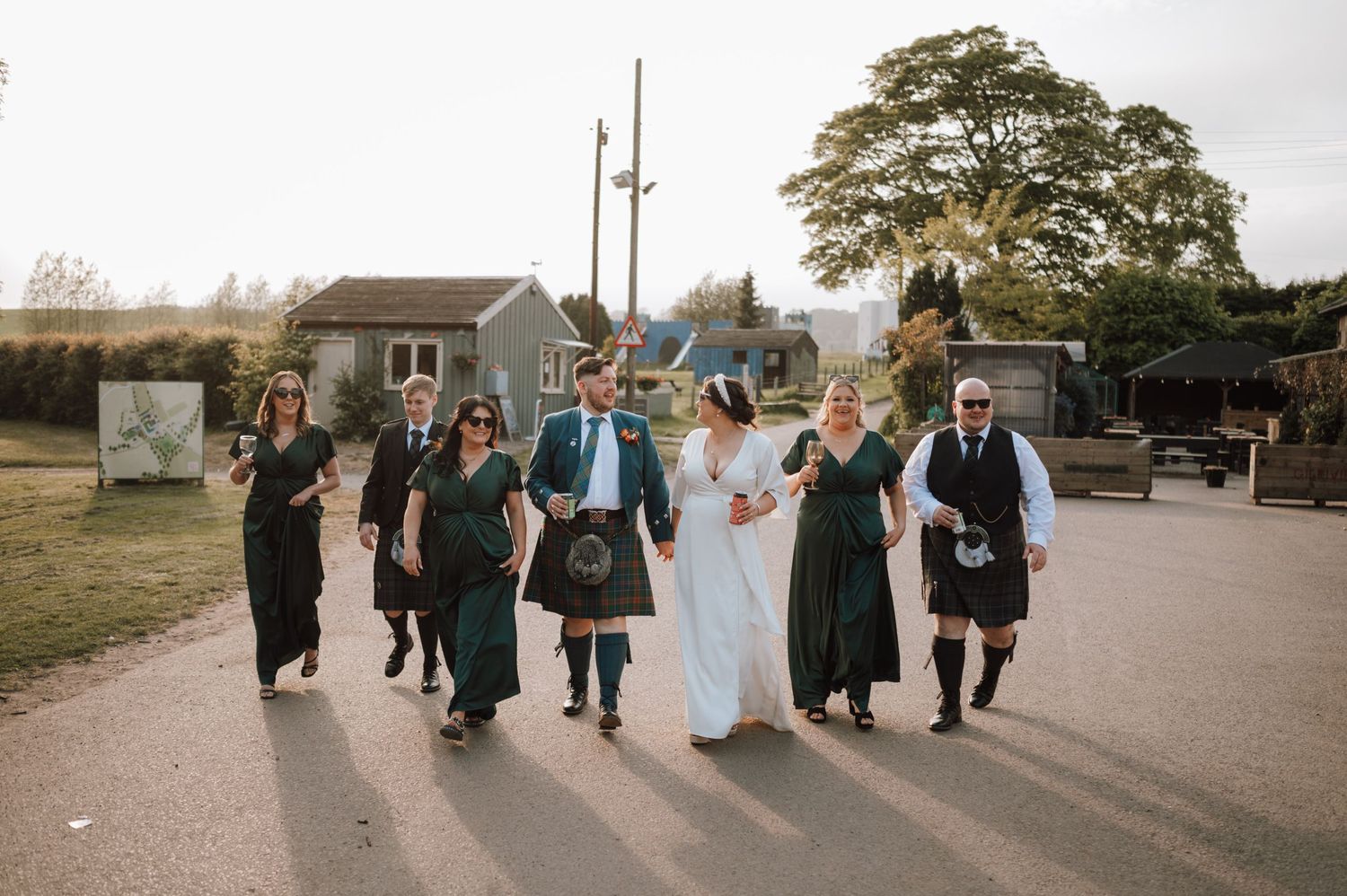 A wedding party walks together outdoors wearing traditional Scottish kilts and formal attire.