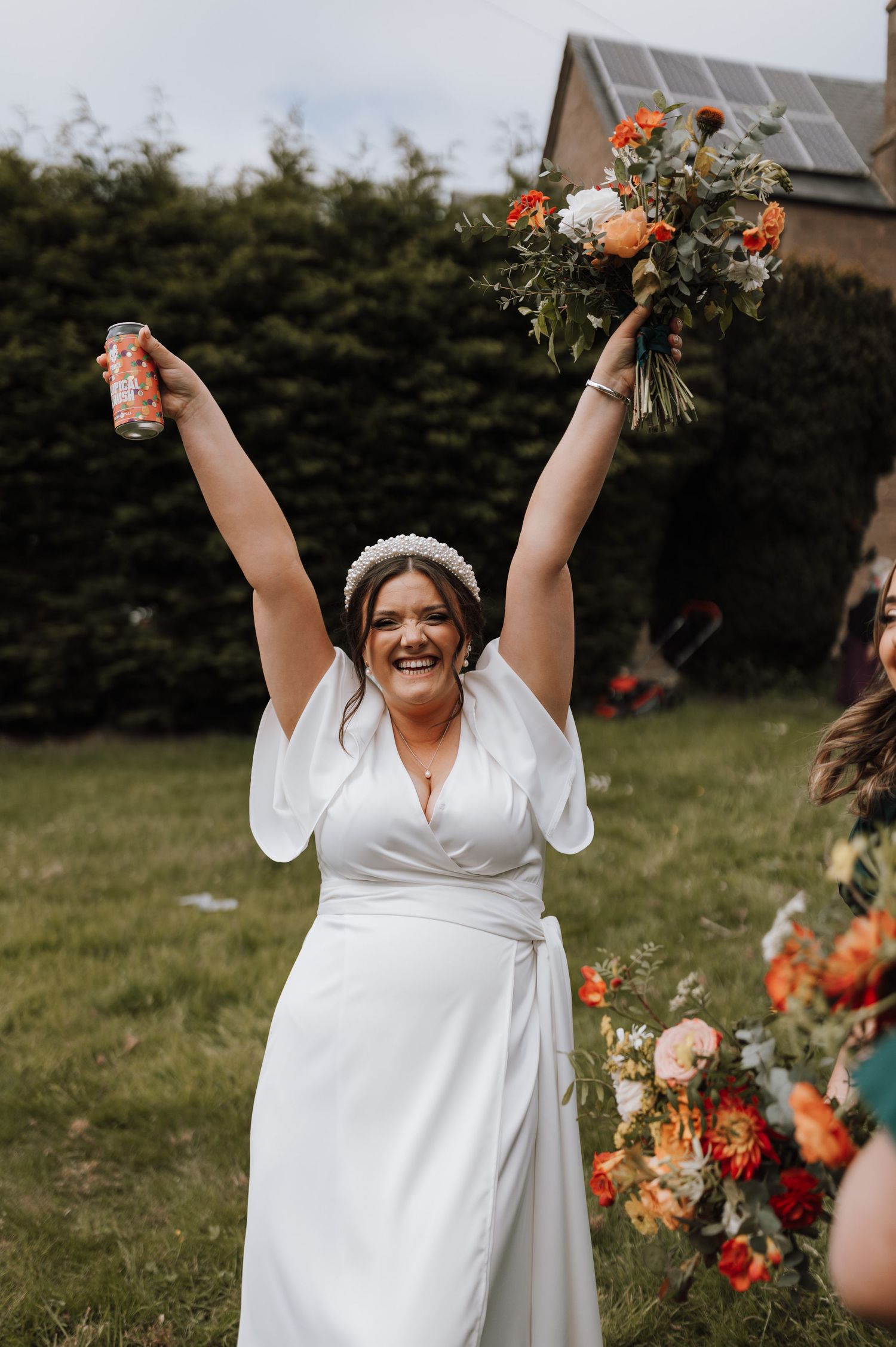 Joyful bride in white dress raises bouquet and drink in celebration outdoors.