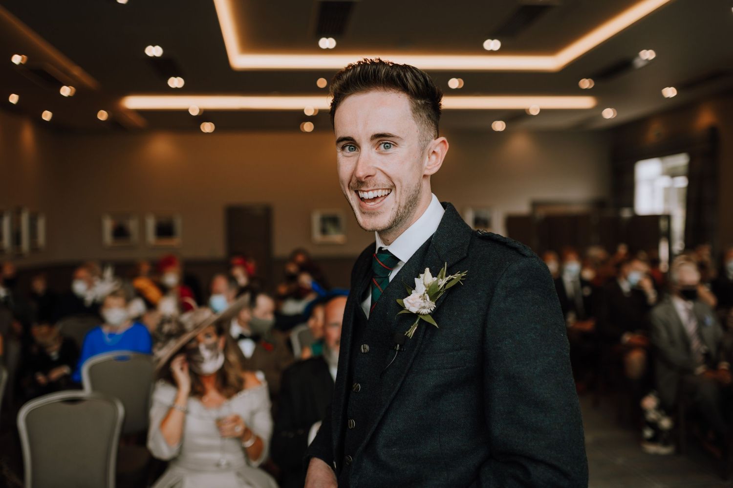 Groom in black suit with boutonniere smiles during indoor wedding ceremony.