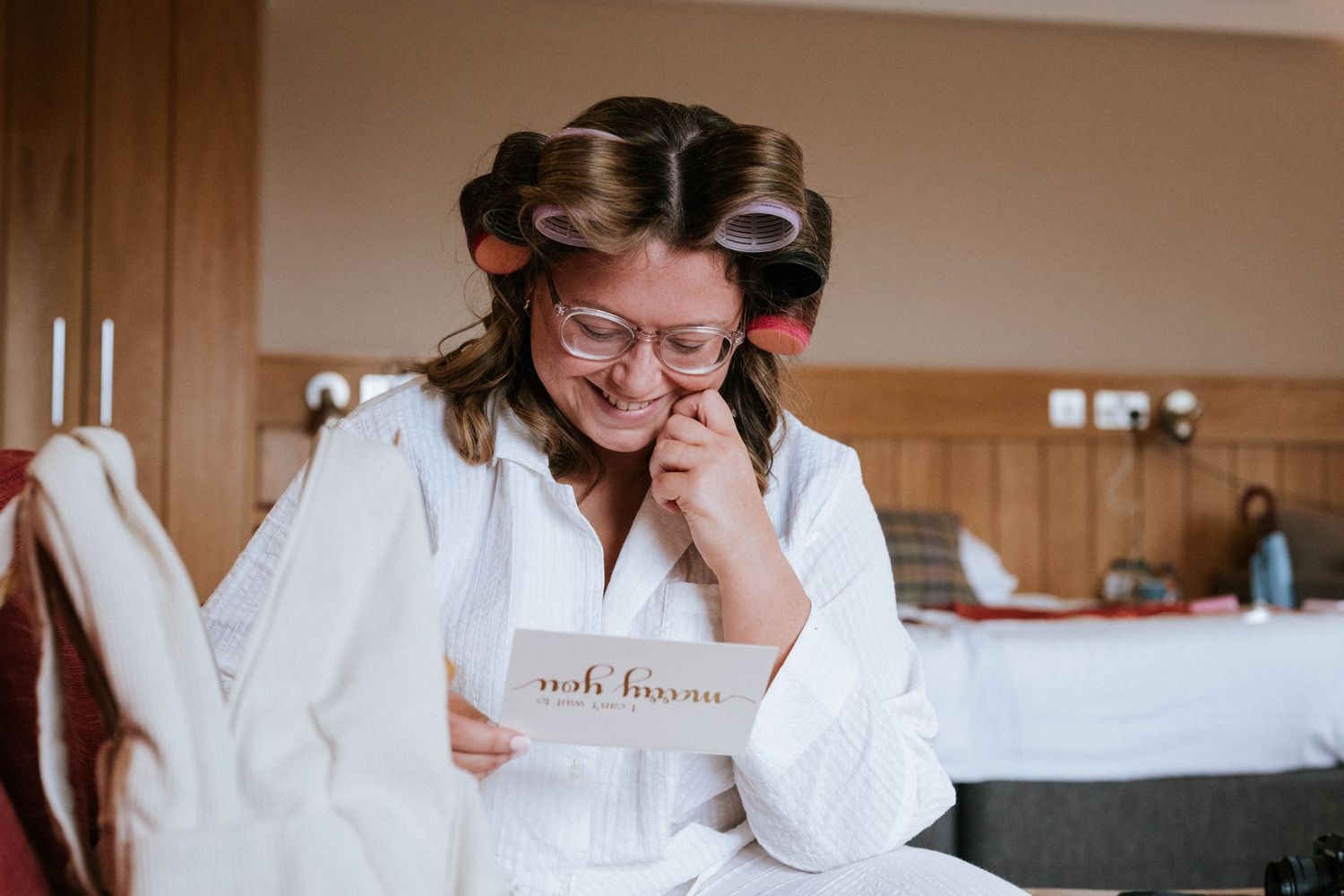 Someone in a white robe reads a note while sitting on a bed during wedding preparations.