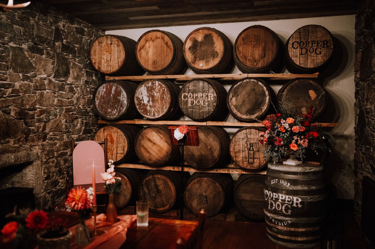 Rustic wine cellar with stacked wooden barrels against a stone wall and red floral decorations.