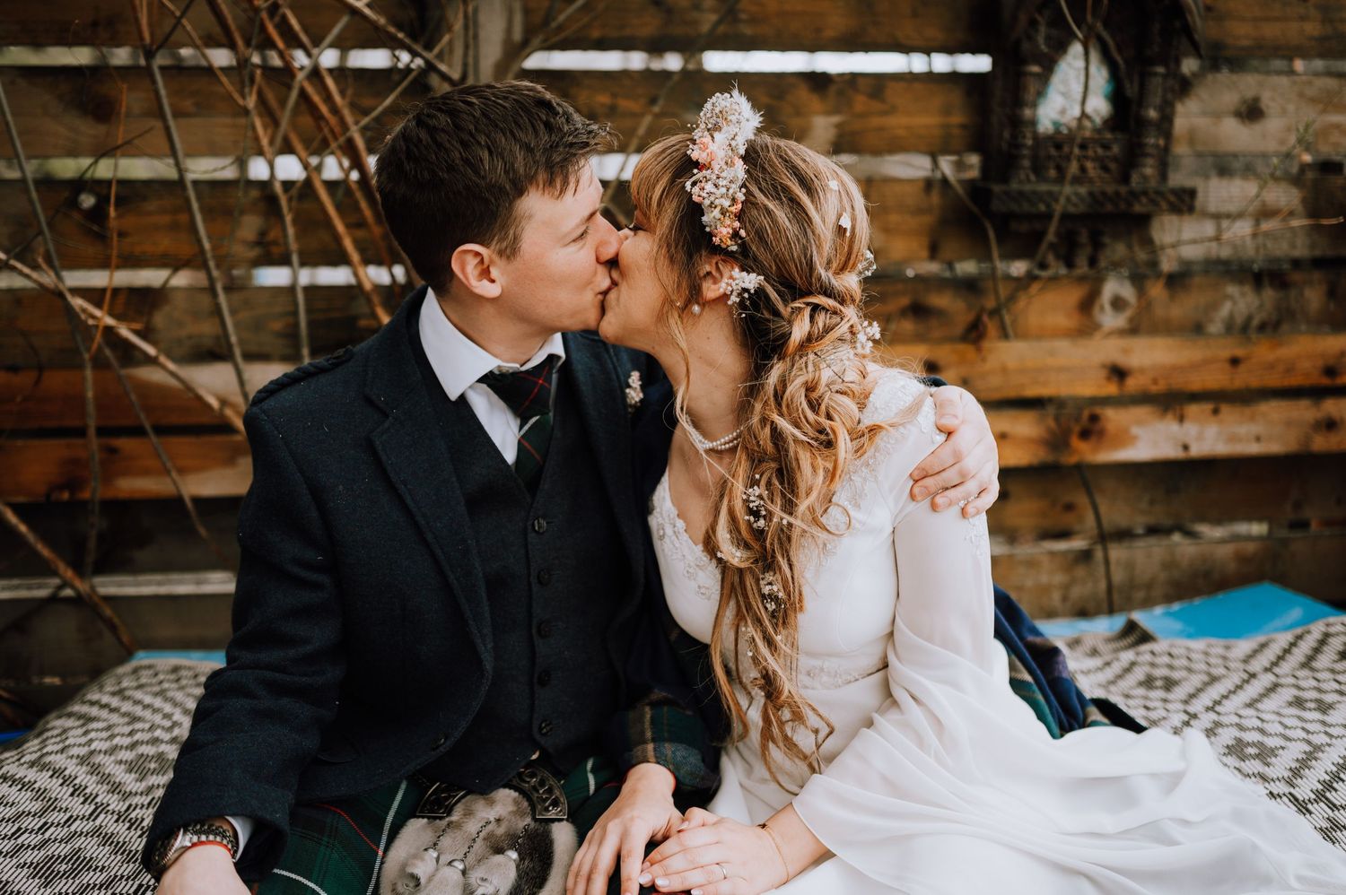 A tender moment between newlyweds in traditional Scottish attire against a rustic wooden backdrop.