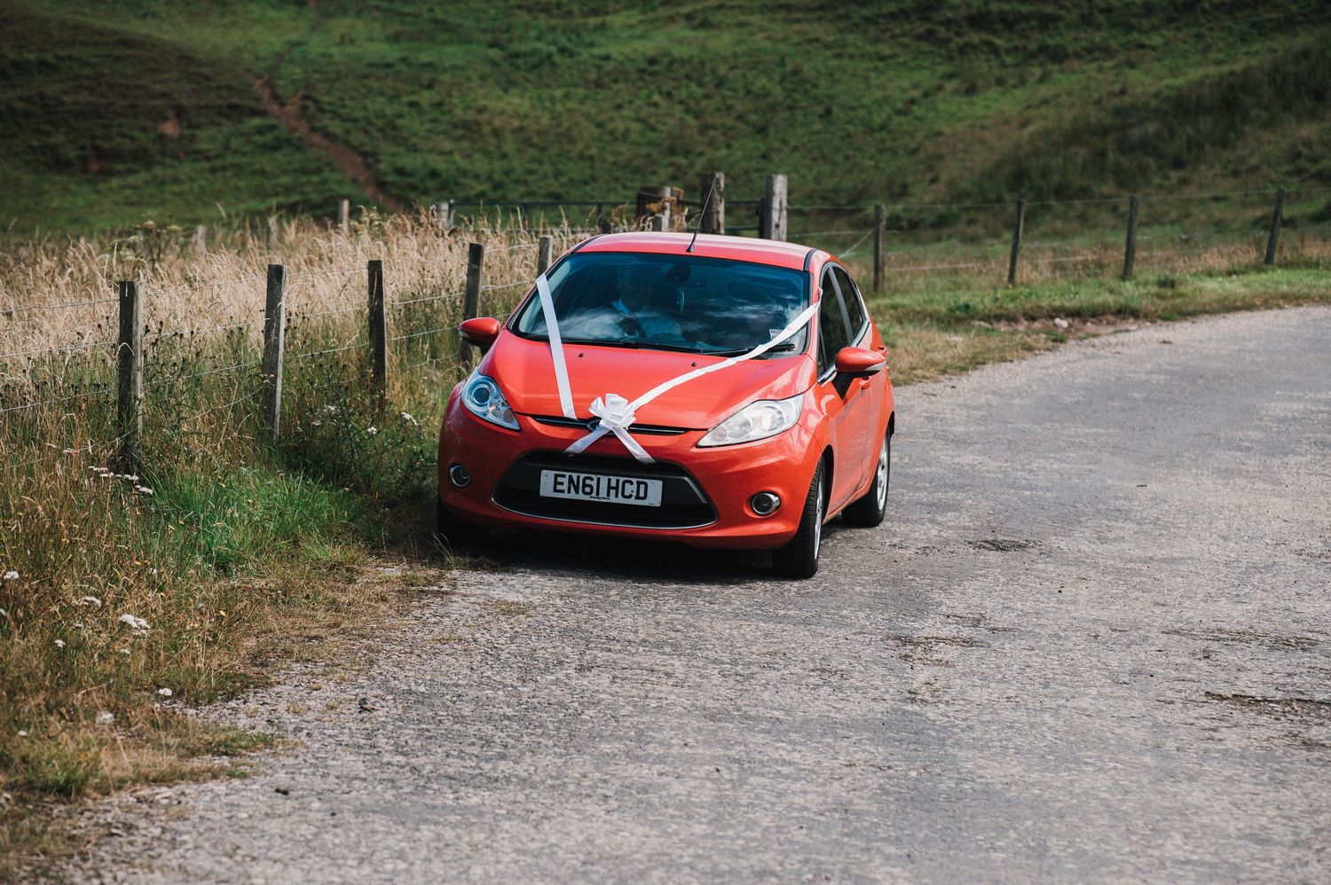 Red car decorated with white ribbons drives along a rural road.