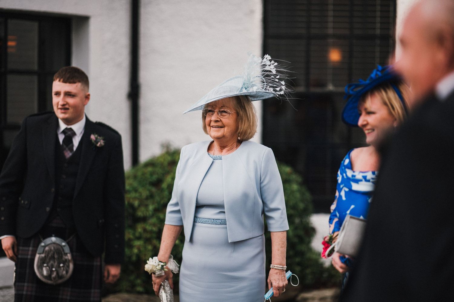 Wedding guests in formal attire and elegant hats gather outside a white building.