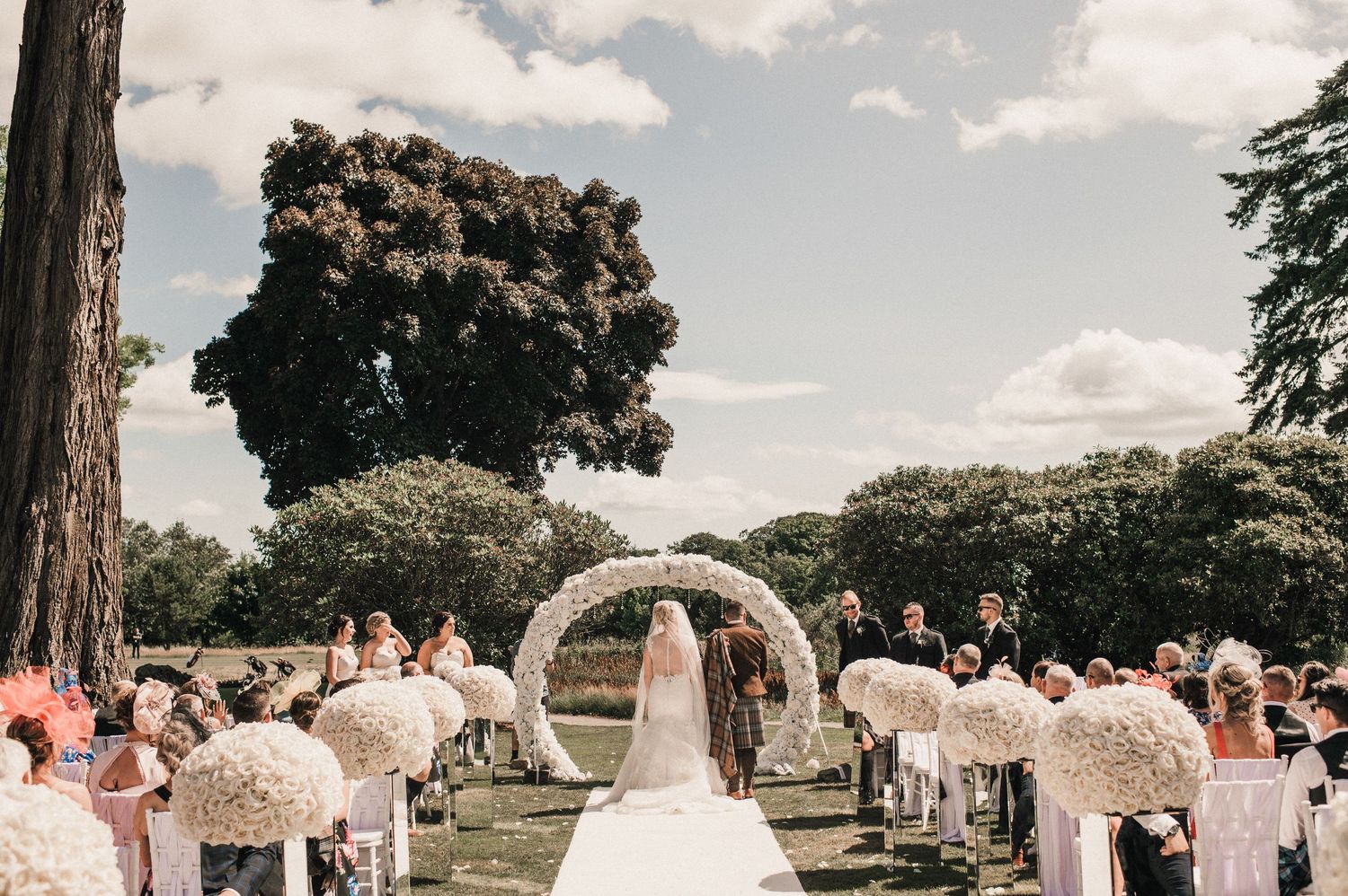 Wedding ceremony viewed from behind with white floral arch and baby's breath decorations.