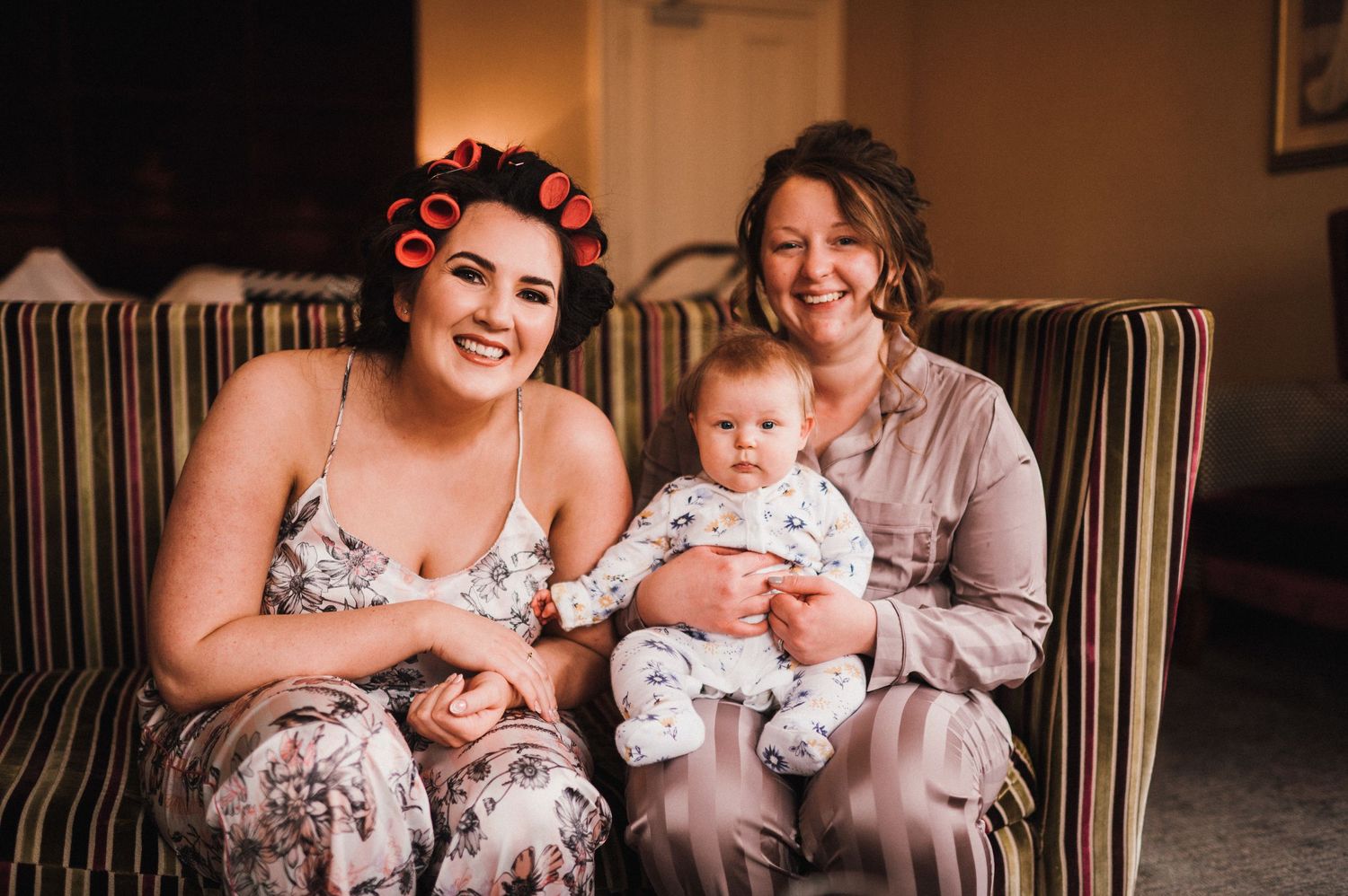 Three people sitting together on striped couch holding baby in white outfit.