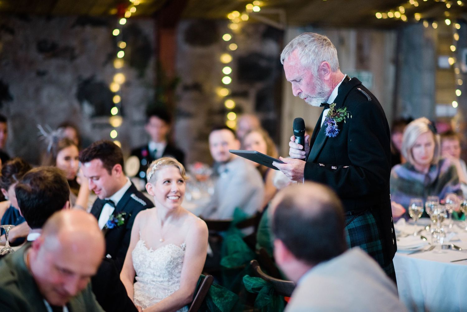 Wedding guests enjoy speeches during a reception in a rustic venue with string lights.