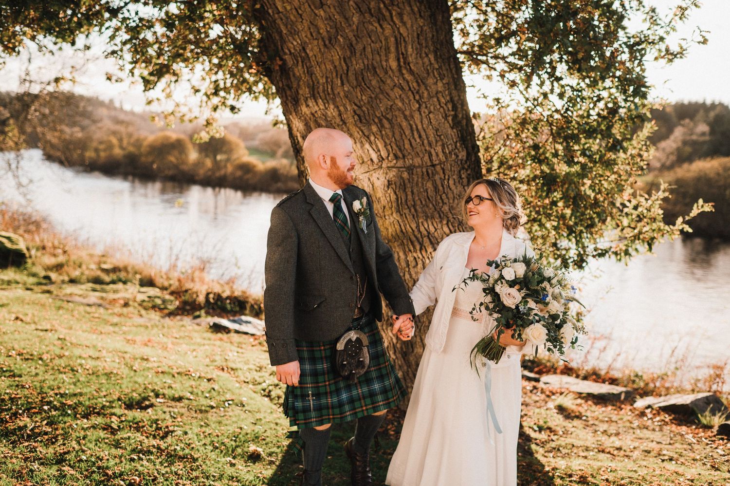 Scottish wedding couple standing by a tree near a river at sunset.