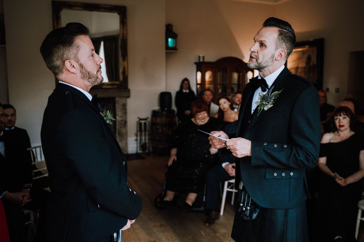 Logie House Wedding Photography. Two people in dark suits embrace during a wedding celebration with guests in the background.