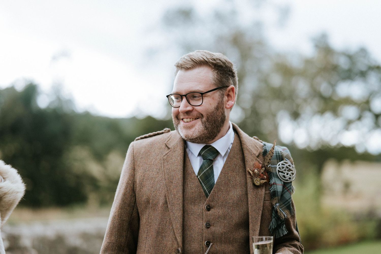 Groom in brown tweed suit and glasses smiling outdoors.