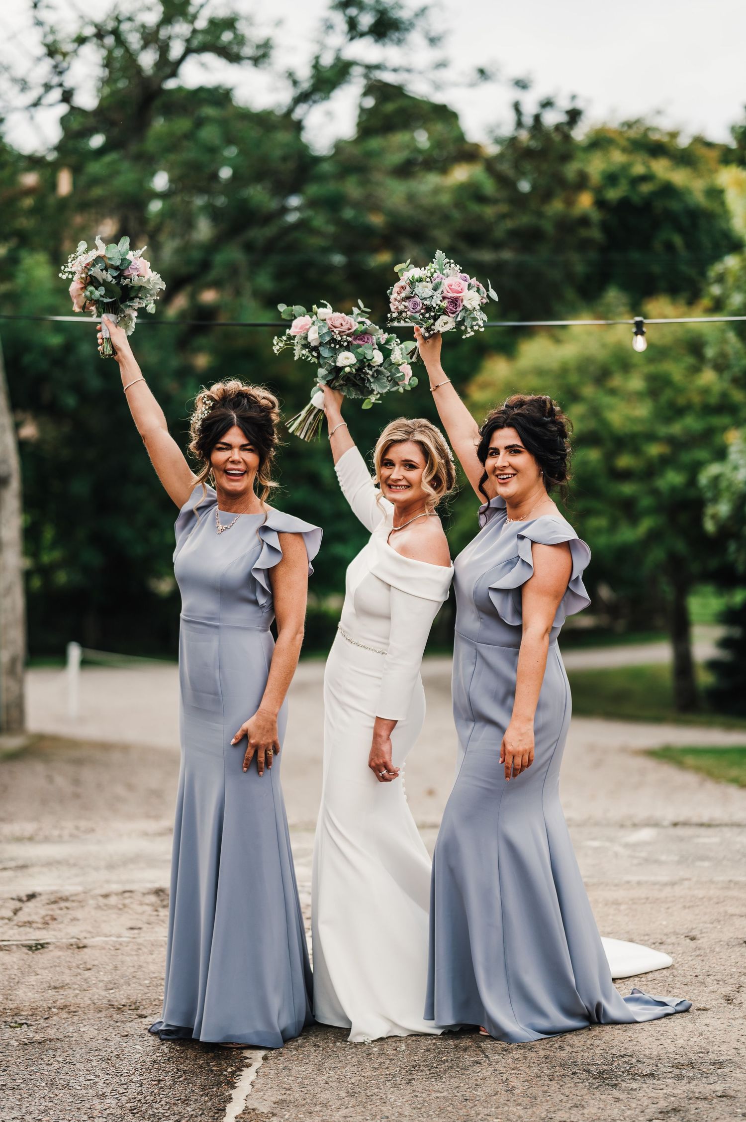Wedding party in grey dresses and white gown celebrating joyfully outdoors with raised bouquets.