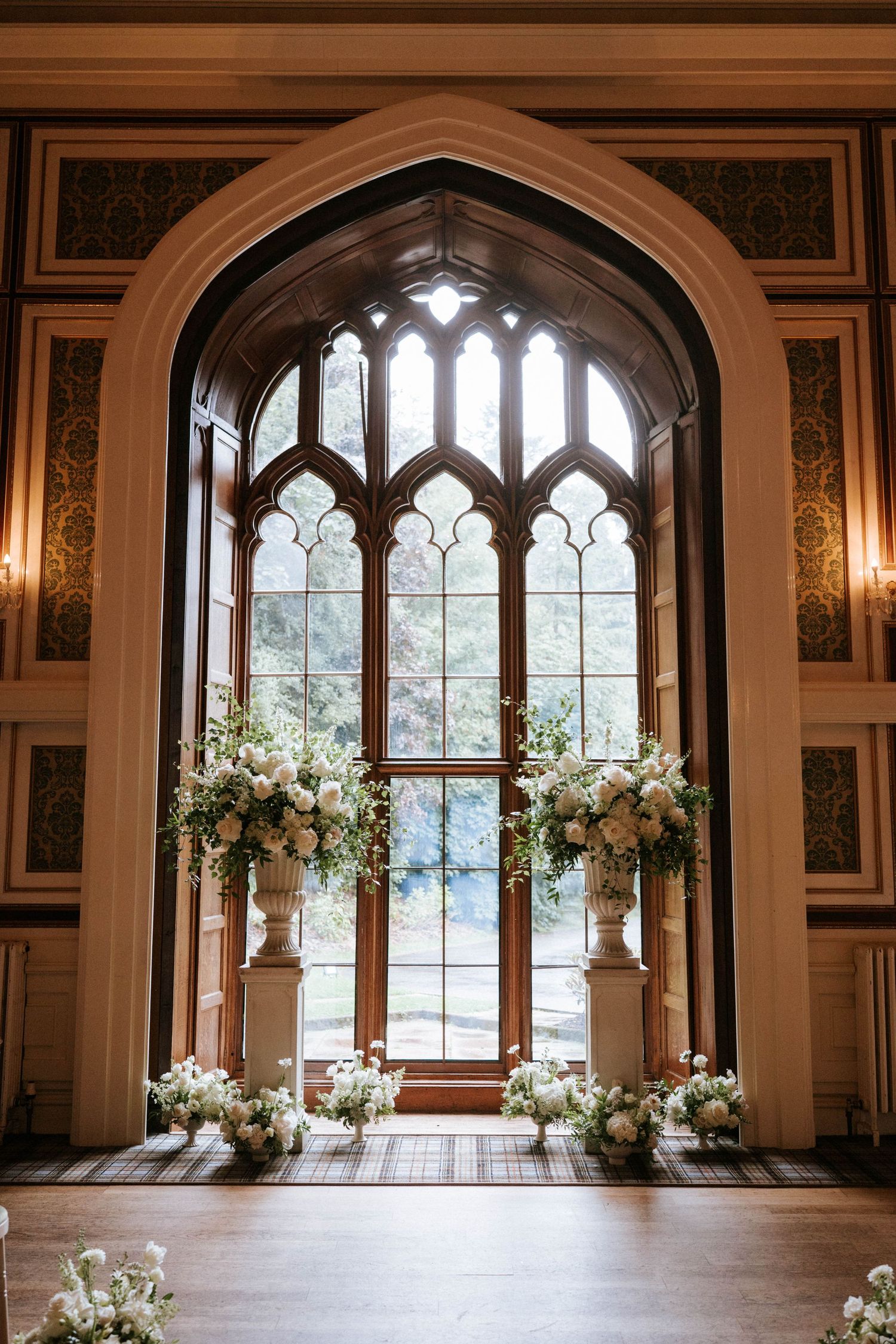 Gothic arched window with ornate details framed by white floral arrangements in a grand wedding ceremony space.