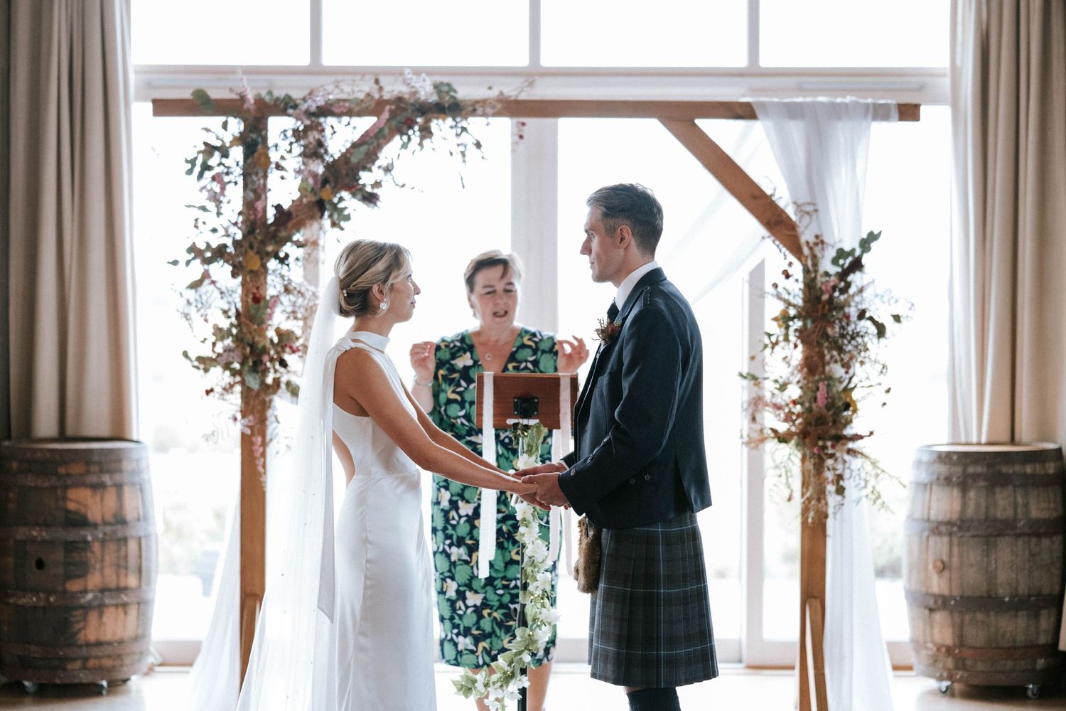 Wedding ceremony under wooden arch decorated with greenery and white curtains.