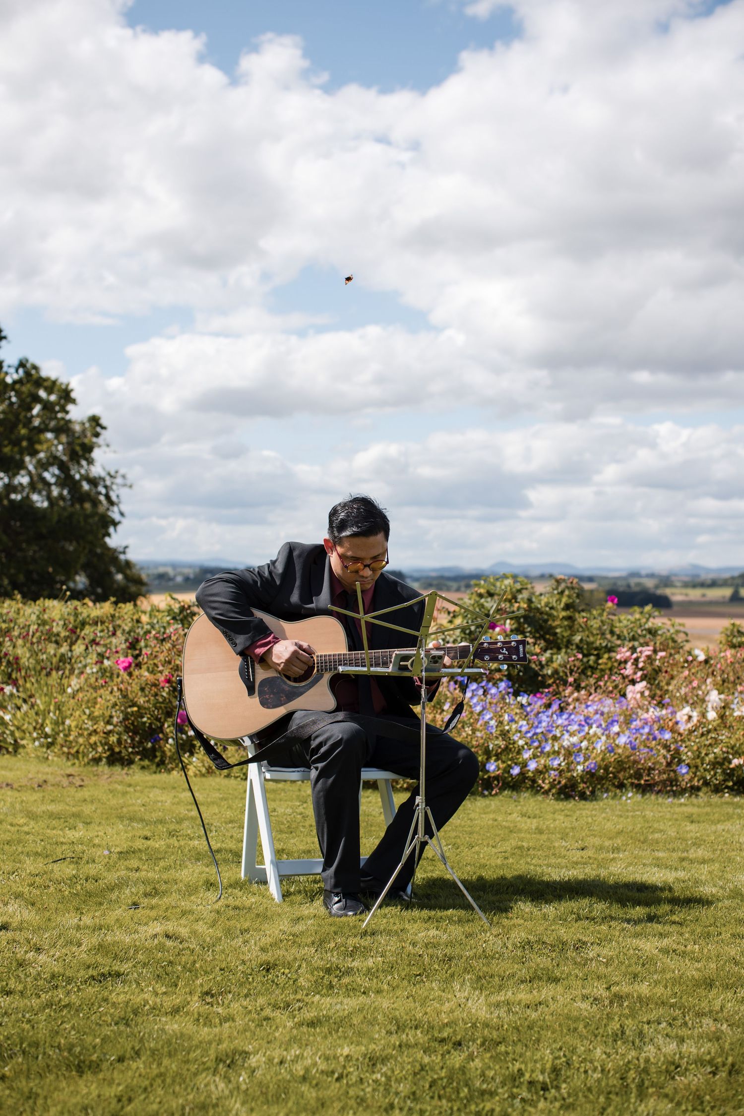Musician performing with acoustic guitar outdoors on a sunny day with cloudy sky.