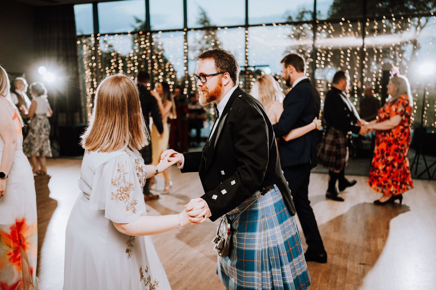Couples dance at a wedding reception with fairy lights decorating the windows.