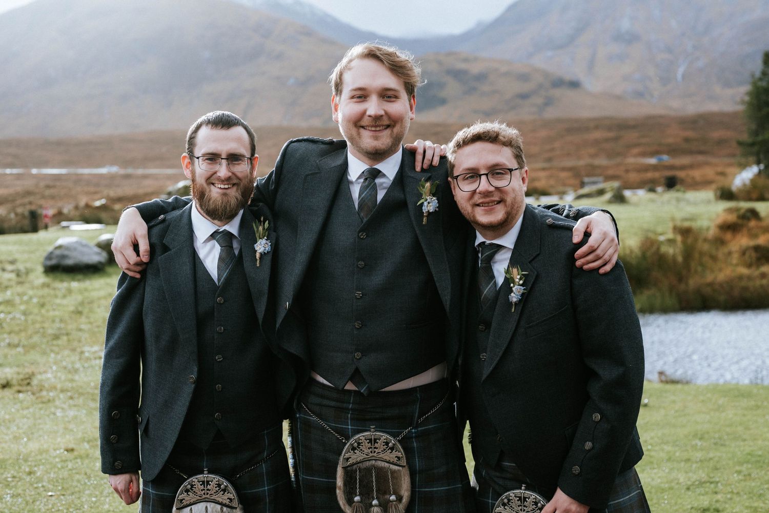 Group wearing traditional Scottish formal wear pose in front of Highland landscape.